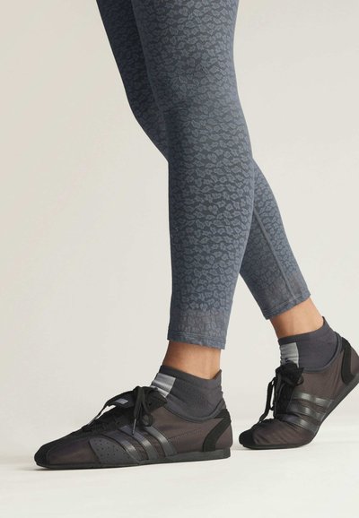 Black athletic shoes with grey stripes, featuring a mesh upper. Worn with dark patterned leggings and grey crew socks, showcasing a sleek fit.