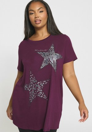 Frau in einem dunkelvioletten kurzärmligen Shirt mit zwei großen Tiermotivsternen und kleinen Verzierungen auf der Vorderseite.