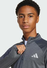 Šedý sportovní pulovr se zipovým límcem, dlouhými rukávy a otvory na palce, doplněný reflexním logem na hrudi.