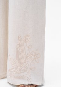 Beige linnen broek met een gestructureerde stof en ingewikkelde lichtroze borduursels van abstracte vormen en bloemen op het onderbeen.