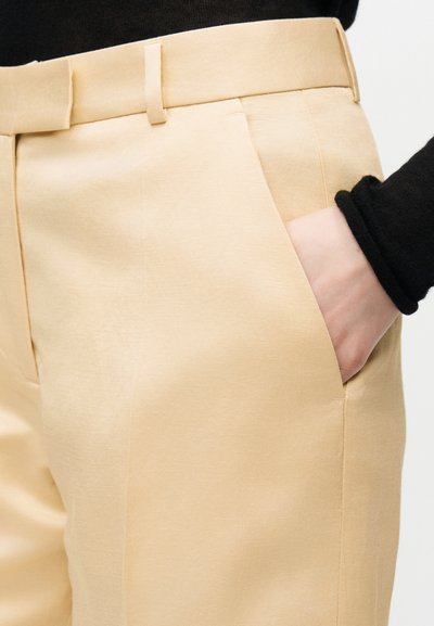 Beige bukser med sidelomme, hånd inni lommen, og svart langermet genser synlig ved håndledd.