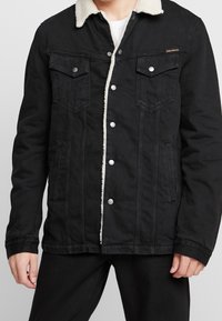 Svart denimjacka med fuskpälskrage, tryckknappar, två bröstfickor och sidfickor; har synlig sömnadsdetalj.