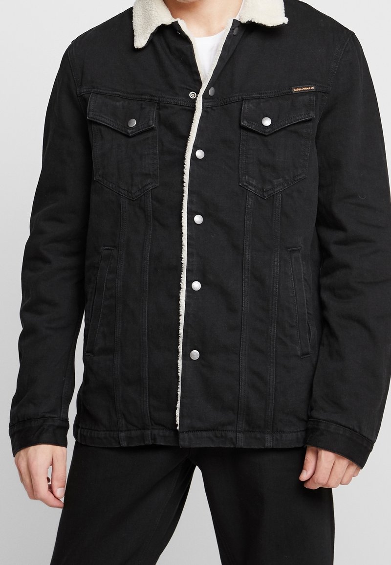 Svart denimjacka med fuskpälskrage, tryckknappar, två bröstfickor och sidfickor; har synlig sömnadsdetalj.