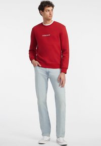 Rød genser med rund hals, med hvit tekst "GUESS JEANS". Kombinert med lyseblå jeans og hvite sneakers, glatt tekstur.