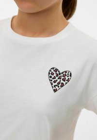 T-shirt en coton blanc arborant un motif de cœur imprimé léopard en noir, rose et blanc sur la partie gauche de la poitrine. Encolure ronde.