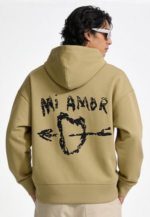 Homme portant un sweat à capuche beige avec un graphisme noir d'un cœur transpercé par une flèche et le texte "Mi Amor" dans le dos, de profil avec les mains dans les poches.