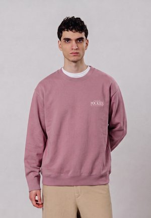 Junger Mann mit kurzen lockigen Haaren trägt ein staubiges rosa Sweatshirt mit weißem Text und beige Hose vor einem schlichten hellgrauen Hintergrund.