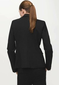 Kobieta z długim brązowym kucykiem, ubrana w czarną marynarkę i pasujące czarne spodnie, stojąca na tle jednolitego jasnego tła, odwrócona tyłem.