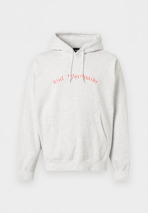 Jeune personne portant un sweat à capuche gris clair avec des patchs graphiques ronds multicolores dans le dos, associée à un pantalon noir, vue de dos.