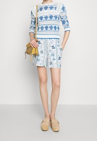 Niebieski i biały sweter z dzianiny z kwiatowym wzorem, w zestawieniu z jasnoniebieskimi spódnico-spodnami w wzory. Kremowe klapki oraz żółta torba na ramię dopełniają całość.