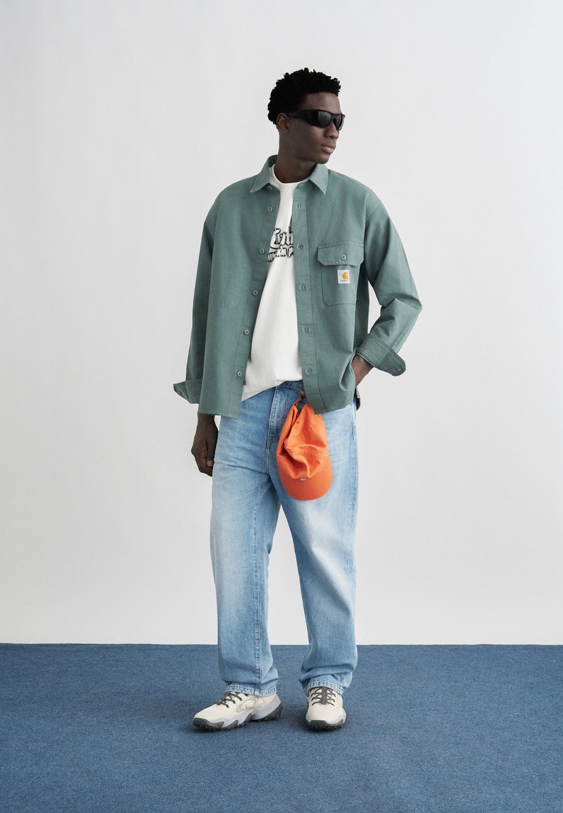 Man wearing sunglasses, green button-up shirt over white graphic tee, light blue jeans, holding an orange cap, standing on blue carpet.