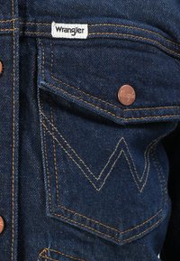 Džínová bunda v tmavomodrej farbe, s bielym označením "Wrangler", mosadznými gombíkmi a kontrastným stehom s charakteristickým cik-cak vzorom na vreckách.