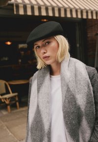 Schwarzes Woll-Baske, grau gemusterter Schal über einem weißen Hemd, strukturierter grauer Blazer; die Person blickt nach vorne vor einem Café-Hintergrund.