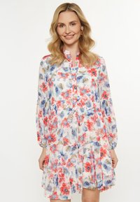 Donna con lunghi capelli ondulati sorride indossando un vestito floreale colorato con maniche lunghe e cintura annodabile, su uno sfondo bianco.