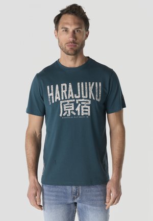 Hombre con camiseta turquesa con texto desgastado "HARAJUKU" y caracteres japoneses, combinada con vaqueros azul claro, de pie frente a un fondo liso.
