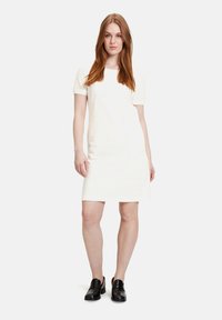 Weißes Kurzarmkleid aus strukturiertem Stoff mit gerader Silhouette, getragen mit schwarzen Slippern, steht vor einem schlichten Hintergrund.