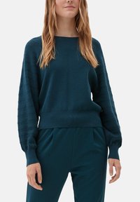 Kobieta ubrana w turkusowy sweter dziergany z teksturowanymi bufkami oraz pasujące turkusowe spodnie na jednolitym tle.