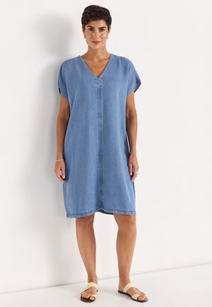 Frau steht und trägt ein locker sitzendes, knielanges blaues Denimkleid mit kurzen Ärmeln und beige flache Sandalen.