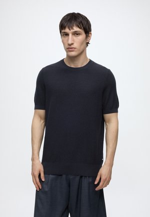 Jeune homme aux cheveux courts et foncés portant un pull en maille noir à manches courtes et un pantalon ample foncé sur un fond clair uni.