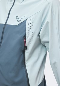 Veste bleu clair et gris foncé avec des points ventilés verticaux et une poche zippée dotée d'une languette rose sur la zone de la poitrine.
