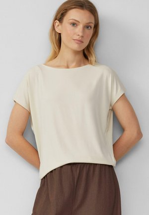 Vrouw met blond haar, gekleed in een crèmekleurig shirt met korte mouwen en losse bruine broek, staande tegen een effen lichtgrijze muur.