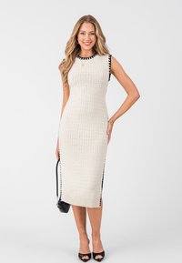 Abito senza maniche in maglia di un beige chiaro con rifiniture nere e spacchi laterali. Presenta un motivo testurizzato e un orlo a costine. Abbinato a tacchi neri.