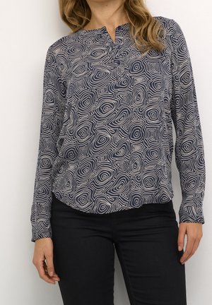 Femme portant un chemisier à manches longues noir et blanc avec des formes concyrtiques abstraites et un pantalon noir, debout devant un fond uni.