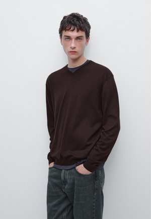 Bruine v-hals trui van een zachte stof, gecombineerd met een donkerblauw shirt eronder. Model poseert met handen in de zakken en draagt donkere jeans.