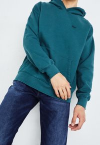 Turkusowa bluza z kapturem wykonana z mieszanki bawełny, z przednią kieszenią kangurką i ściągaczami, zestawiona z niebieskimi dżinsami.