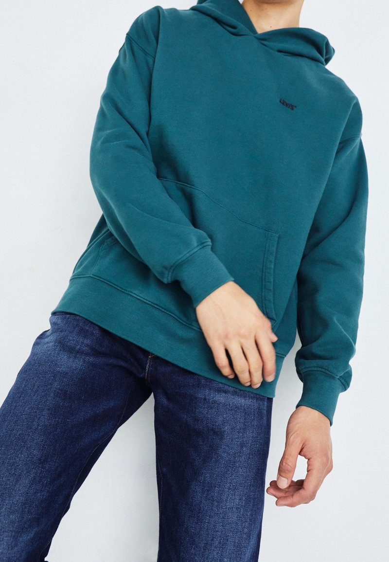 Turkusowa bluza z kapturem wykonana z mieszanki bawełny, z przednią kieszenią kangurką i ściągaczami, zestawiona z niebieskimi dżinsami.
