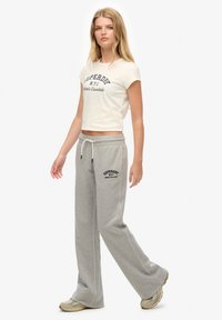 T-shirt courte beige clair avec lettrage bleu marine, associée à un jogging gris arborant un logo noir. Les deux sont confectionnés dans un tissu en coton doux.
