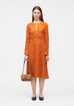 Vrouw in een oranje midi-jurk met lange mouwen en subtiele patronen, draagt een beige handtas met een gouden kettingriem en zwarte platte schoenen.