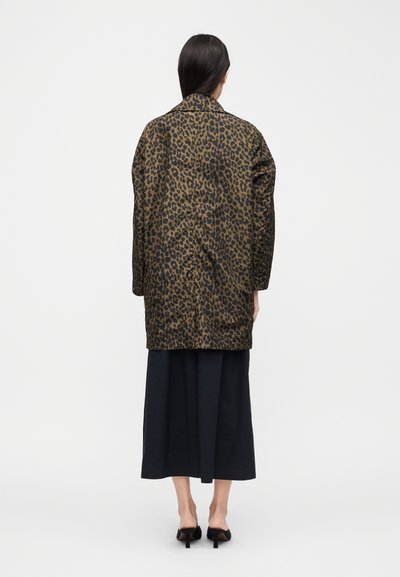 Γυναίκα με μακριά σκούρα μαλλιά, φορώντας παλτό μέχρι το γόνατο με λεοπάρ σχέδιο, μαύρο παντελόνι φαρδιάς γραμμής και μαύρα mules με τακούνι, που στέκεται με την πλάτη προς τα εμάς.