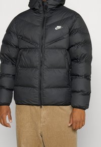 Black puffer jacket with vertical and horizontal stitching, zip closure, side pockets, and hood, detailed with a small white logo on the chest.