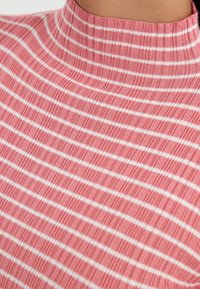 Růžová ribková tkanina s bílými pruhy, která má design s vysokým krkem. Textura je měkká a pružná, zvýrazňuje vzor.