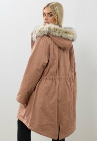 Ruskea parka-takki, jossa on karvalla vuorattu huppu, elastinen vyötärö, sivutaskut ja takaventti. Kangas on sileä ja hieman kiiltävä.