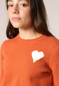 Pull en tricot orange avec un col rond et un détail en forme de cœur blanc duveteux sur la partie supérieure gauche de la poitrine. Texture douce.