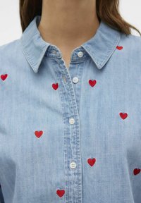 Camicia in denim azzurro chiaro con ricami a forma di cuore rossi sparsi sul davanti, colletto classico e chiusura con bottoni bianchi.