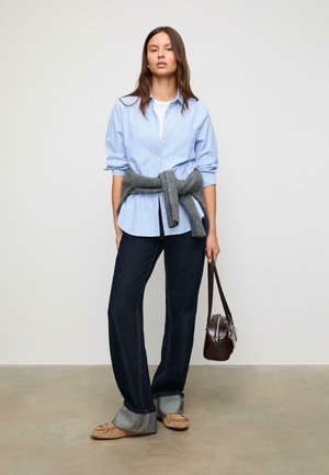 Donna in piedi che indossa una camicia a righe blu, jeans scuri con risvolti, maglione grigio legato in vita, scarpe beige, che tiene una borsa marrone.