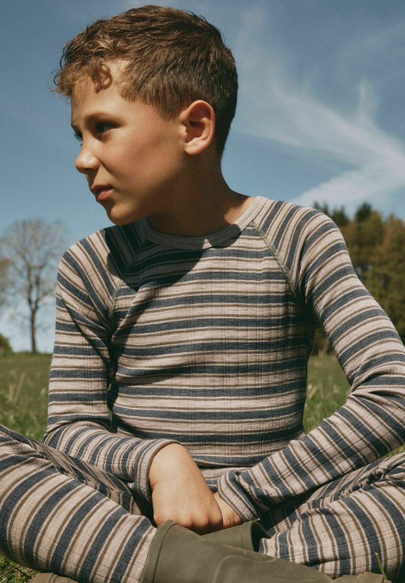 Garçon aux cheveux bouclés portant un t-shirt à manches longues rayé et un pantalon, assis en tailleur dans un champ herbeux sous un ciel bleu regardant sur le côté.