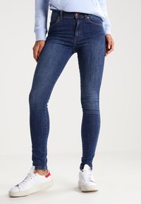 Jeans ajustados de color azul oscuro hechos de denim con una textura suave, que cuentan con una cintura alta y un diseño estándar de cinco bolsillos. Zapatillas blancas con detalles rojos.