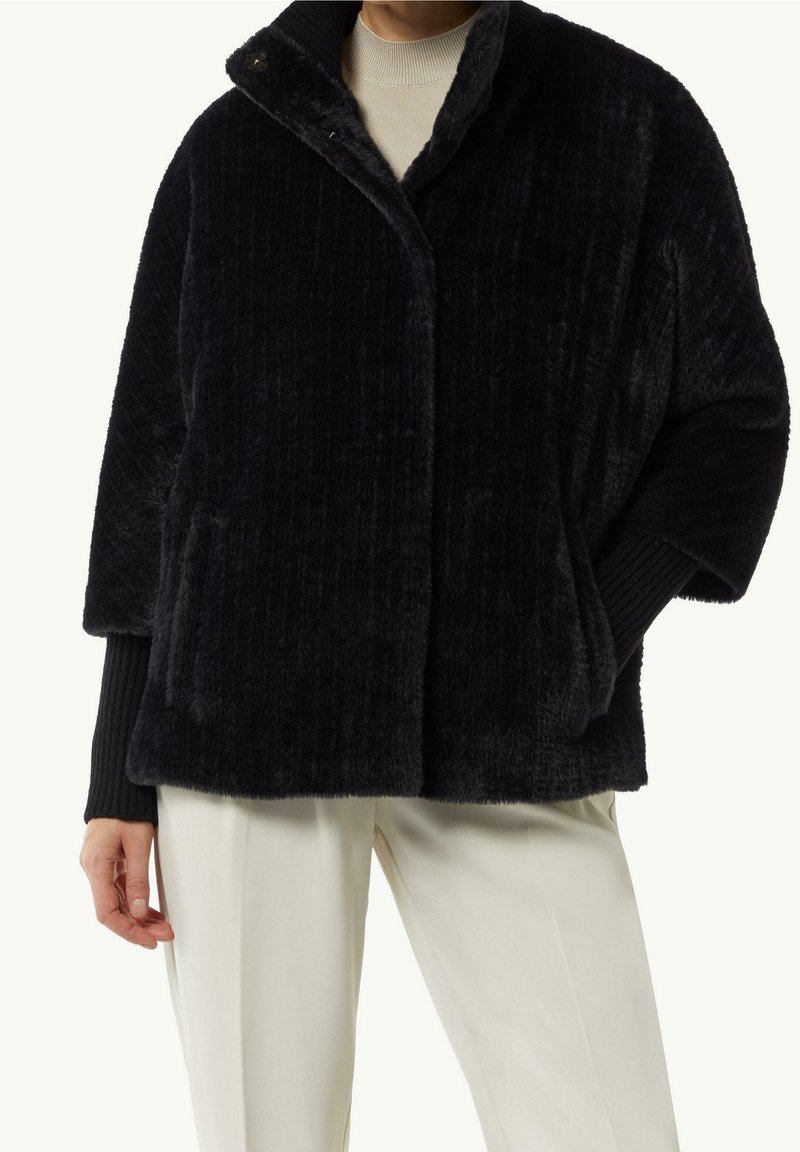 Persoon in een zwart pluche jack met geribde manchetten, een beige top en gebroken witte broek, staande met één hand in de jaszak.