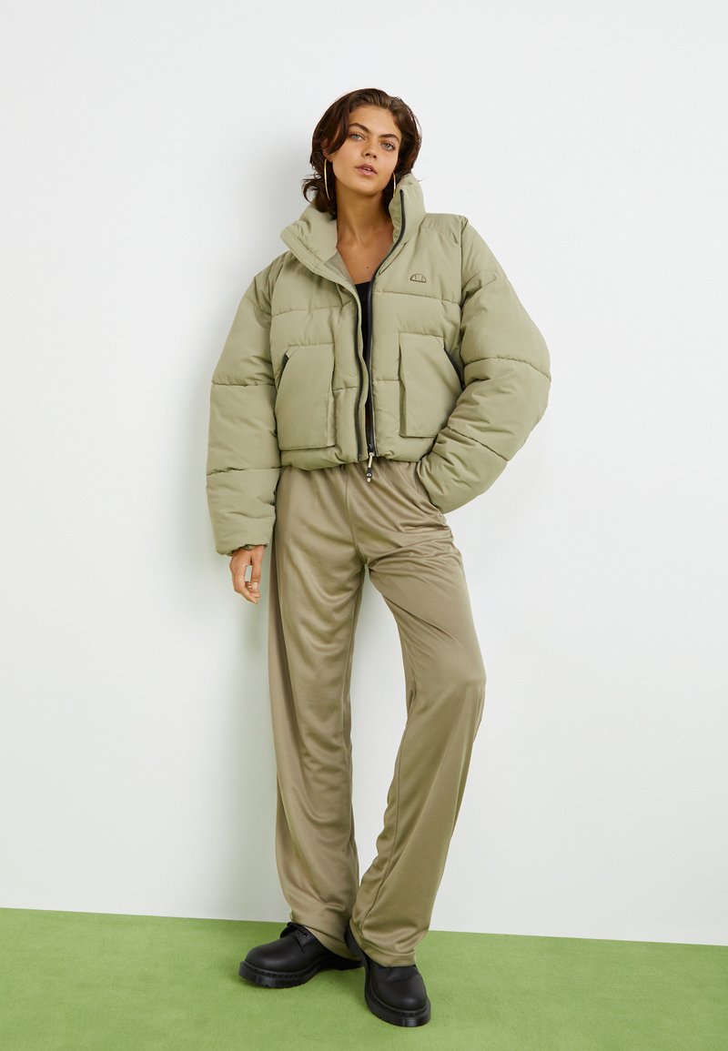 Puchowy oliwkowozielony krótki kurczak z dwoma przednimi kieszeniami i zapięciem na zamek, w połączeniu z beżowymi spodniami i czarnymi butami na jasnym tle.