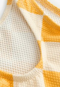 Tissu à carreaux jaune et crème avec une surface texturée. La couche intérieure présente un motif léger en maille. Coutures visibles sur les bords.