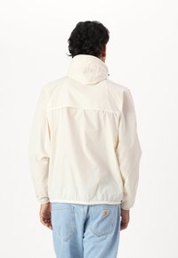 Lehká krémová bunda s kapucí, elastickými manžetami, utěsněným zadním švem a šňůrkou na krku. Kombinována s světle modrými džínami.