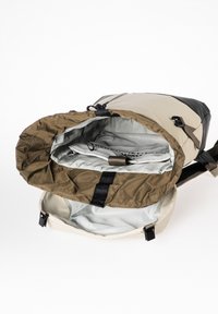 Rucksack mit einem beigen Äußeren und dunklen Akzenten, mit einem braunen Innenfutter, Zugverschluss und einem glatten silbernen Innenraum.