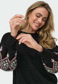 Črna dukserica z vzorčastimi, teksturiranimi rokavi, okrašenimi z belimi, roza in zelenimi cvetličnimi vezeninami. Okrugel ovratnik, sproščen kroj.