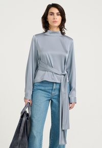 Lysegrå satin bluse med lange ærmer, høj halsudskæring og et bælte i taljen, parret med blå denim. Modellen holder en mørkegrå taske.