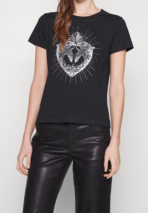 Nainen pukeutuneena mustaan, lyhythihaiseen t-paitaan, jossa valkoinen sydänkuvio kahdella linnulla ja kukkaelementeillä, yhdistettynä mustiin nahkahousuihin.