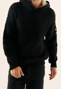 Svart hooded sweatshirt gjort av texturerat stickat tyg, med en kontrasterande fleecefoder, ribbade manschetter och en logotyp-patch på ärmen.